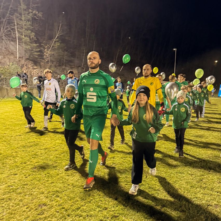 A soccer player leads a group of children in green jerseys on a field, holding hands. Some children hold balloons.