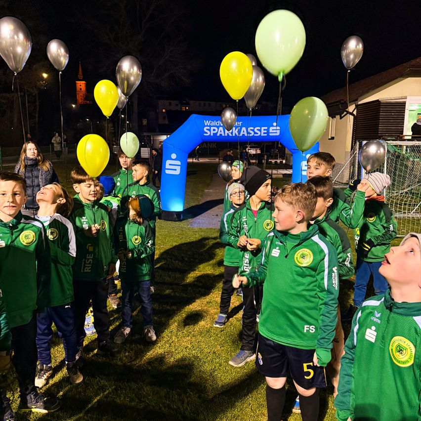 A group of children in green jerseys stands under a blue arch marked with Sparkasse, looking up at yellow balloons.