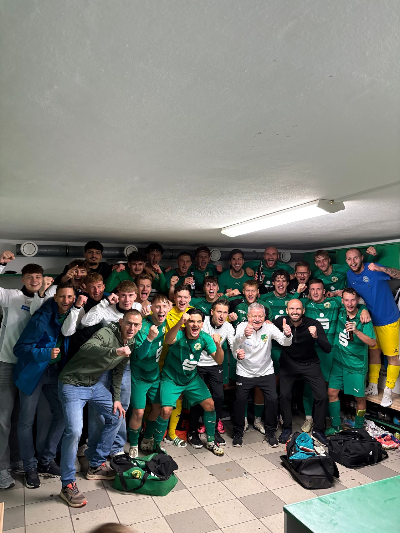 A soccer team celebrates in a locker room, wearing green uniforms, with some holding drinks.