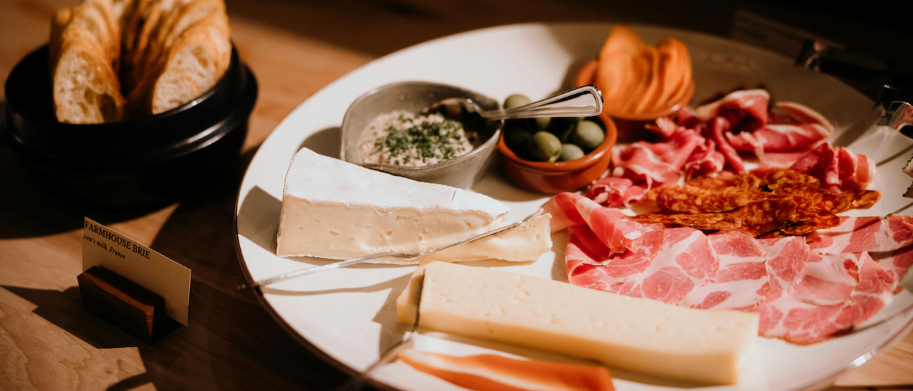 Ein weißer Teller mit verschiedenen Käsesorten, geschnittenem Fleisch, Oliven und einer Dip-Schüssel. Brot befindet sich auf der linken Seite und ein Messer liegt auf dem Teller.
