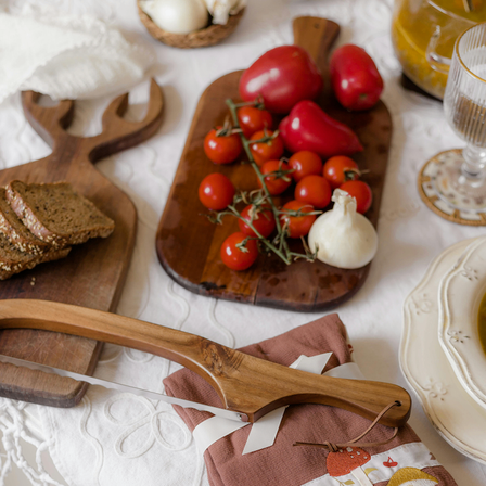 Ein Holzschneidebrett mit roten Tomaten, Knoblauch und einem Messer liegt auf einem Tisch mit weißer Tischdecke. Daneben befinden sich zwei weitere Holzbretter mit Brot und ein Glasbecher.