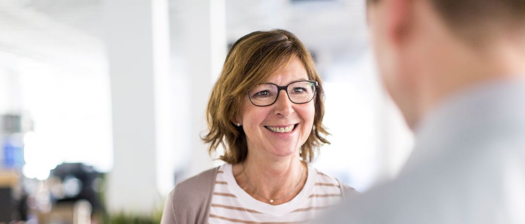 Eine Frau mit Brille lächelt, während sie einen Marker und ein Stück Papier hält, möglicherweise in einem Büro. Ein Mann schaut sie an.