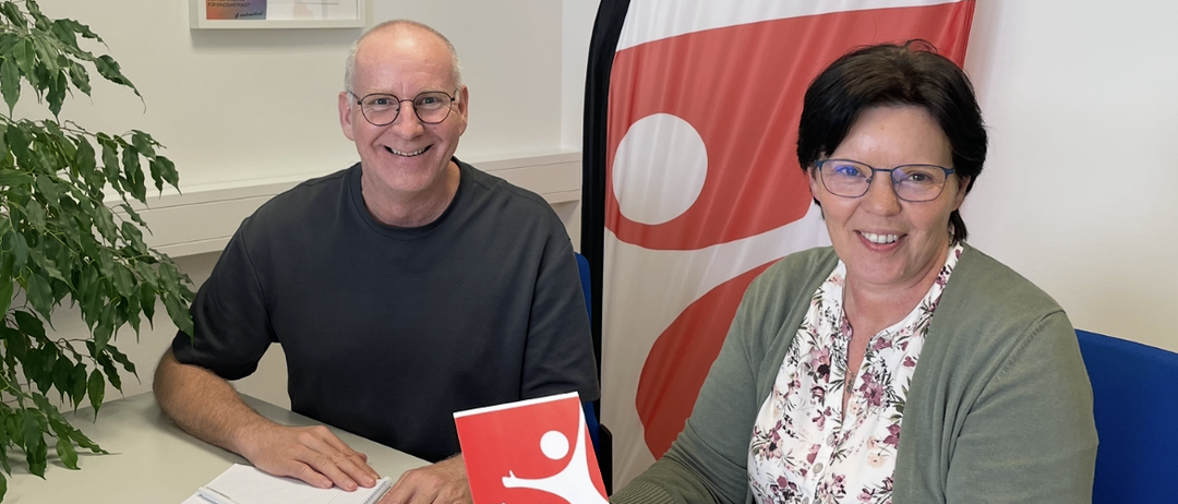 Ein Mann und eine Frau sitzen lächelnd am Tisch, vor ihnen ein Buch und ein Stift. Hinter ihnen eine rot-weiße Fahne.