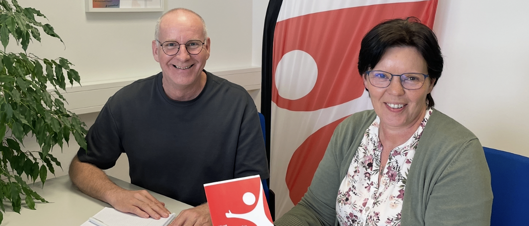 Ein Mann und eine Frau sitzen an einem Tisch mit einem rot-weißen Buch. Der Mann lächelt, während die Frau ein Notizbuch und einen Stift hat. Sie tragen beide eine Brille und eine Uhr. Hinter ihnen ist eine rot-weiße Fahne mit einem Logo zu sehen.