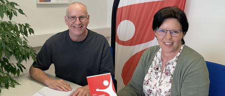 Ein Mann und eine Frau sitzen lächelnd am Tisch, mit einem Buch und einer Fahne im Hintergrund.