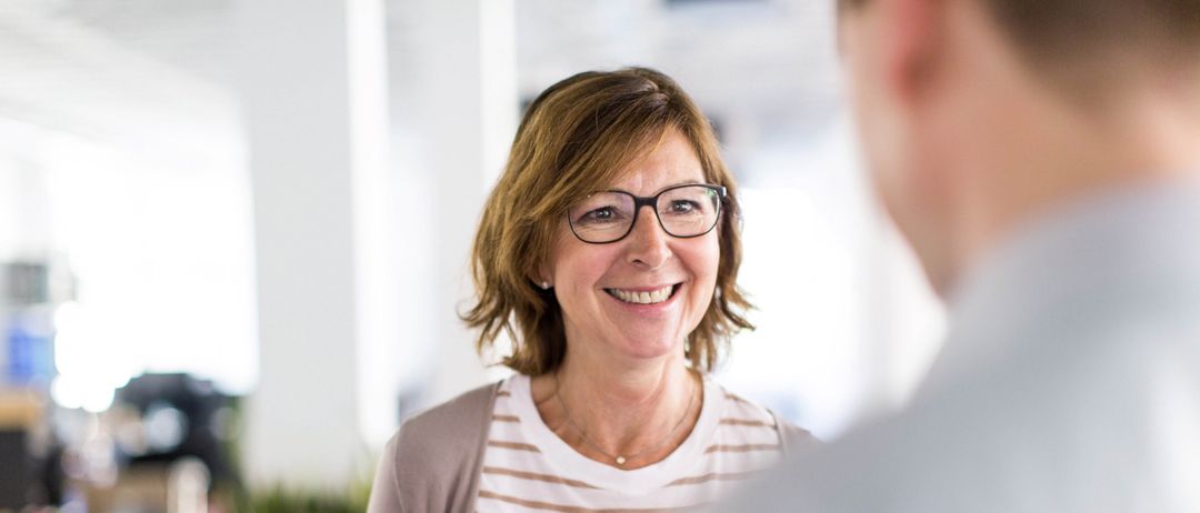 Eine Frau mit Brille und gestreiftem Shirt lächelt einen Mann in einer Büroumgebung an. Sie hält einen grünen Marker und ein Stück Papier.