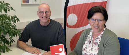 A man and a woman sit at a table, smiling and holding a red booklet. The man wears glasses and a black shirt, while the woman wears glasses and a floral blouse. A notebook and pen are on the table, and a flag with a red and white design is behind them.