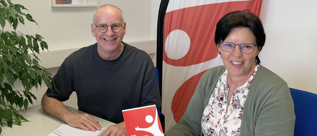 Ein Mann und eine Frau sitzen an einem Schreibtisch, lächeln und halten ein rotes Buch mit dem Titel 'Fit für die Arbeit'. Hinter ihnen ist eine Flagge mit einem roten und weißen Design.