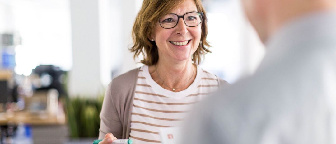 Eine Frau mit Brille und einer Halskette lächelt und hält einen grünen Marker. Sie steht vor einer verschwommenen Person.