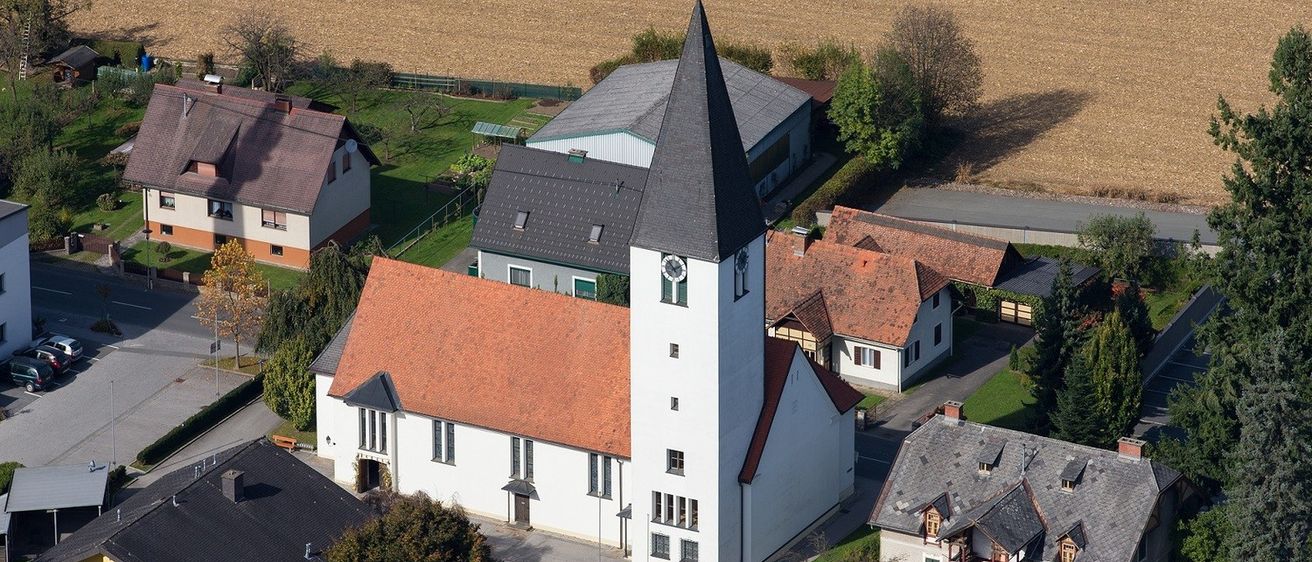 Ein Luftbild einer Kirche mit hohem Turm und rotem Dach. Die Kirche ist von Häusern und einem Bauernhof umgeben, mit einem großen Feld im Hintergrund.