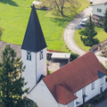 Eine Luftaufnahme einer Kirche mit Turm, rotem Dach und weissem Körper. Es gibt zwei Uhren auf dem Turm.