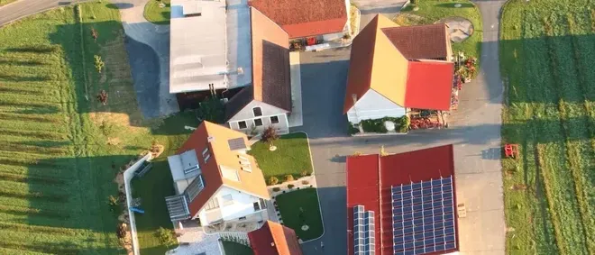 Luftaufnahme einer Vorstadtsiedlung mit mehreren Häusern. Jedes Haus hat ein rotes Dach und Solarpanele. Die Häuser sind von grünem Gras und Einfahrten umgeben.