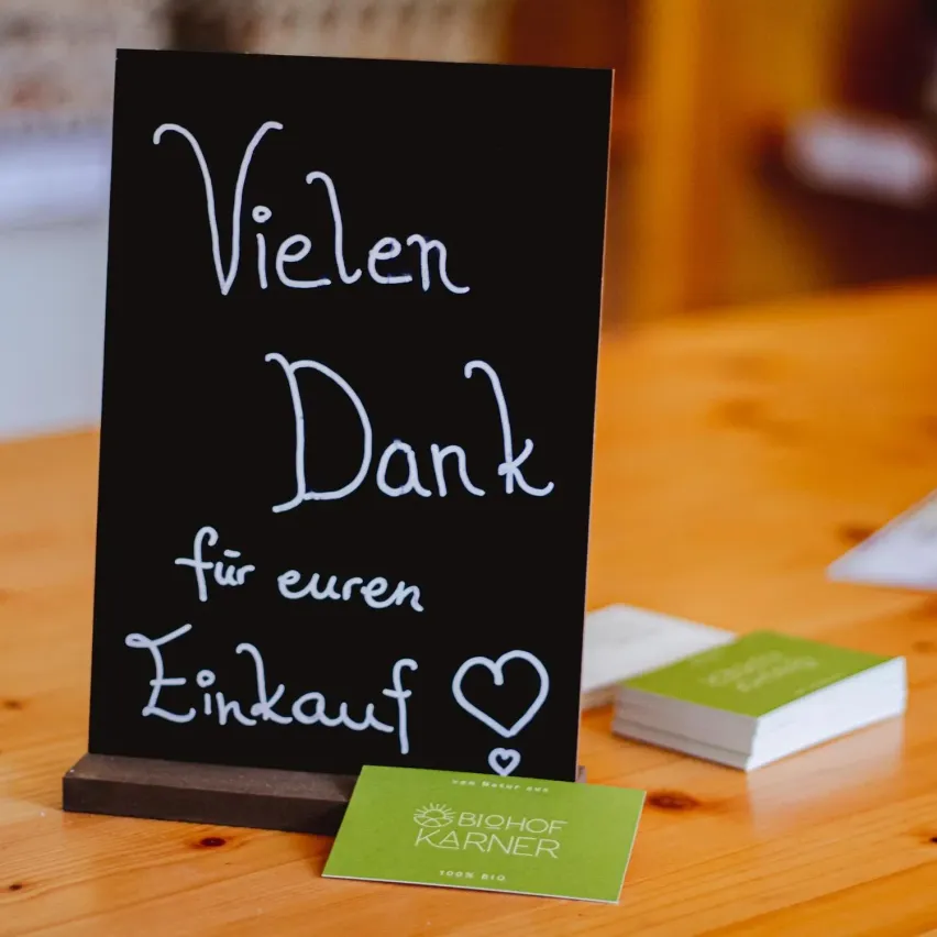 Ein schwarzer Kreidetafel steht auf einem Holztisch mit einer Nachricht, die 'Vielen Dank fur euren Einkauf' lautet. Ein Herzsymbol ist auf der Tafel gezeichnet. Daneben liegt eine grüne Visitenkarte.