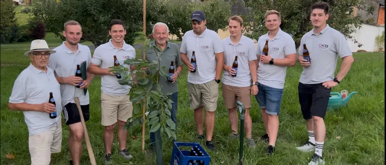 Eine Gruppe von Männern steht draußen, lächelt, mit einem jungen Baum und Bierflaschen. Sie sind in Freizeitkleidung gekleidet und halten Bierflaschen, mit einem Bierkasten, der mit 'PUNTIGAMER' beschriftet ist, davor.
