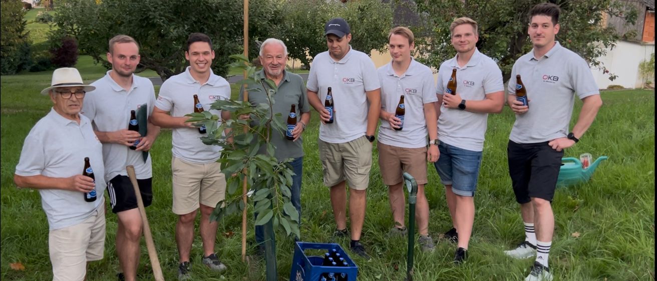 Eine Gruppe von Männern steht draußen, lächelt, mit einem jungen Baum und Bierflaschen. Sie sind in Freizeitkleidung gekleidet und halten Bierflaschen, mit einem Bierkasten, der mit 'PUNTIGAMER' beschriftet ist, davor.