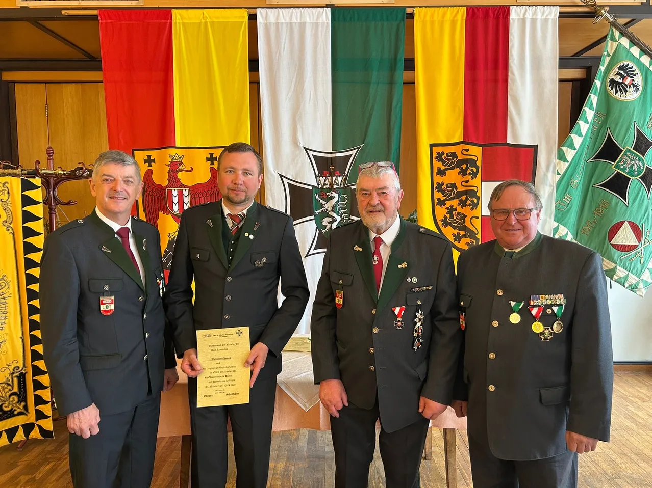 Vier Männer in Uniformen mit Medaillen stehen vor Fahnen. Der Mann in der Mitte hält ein Zertifikat.