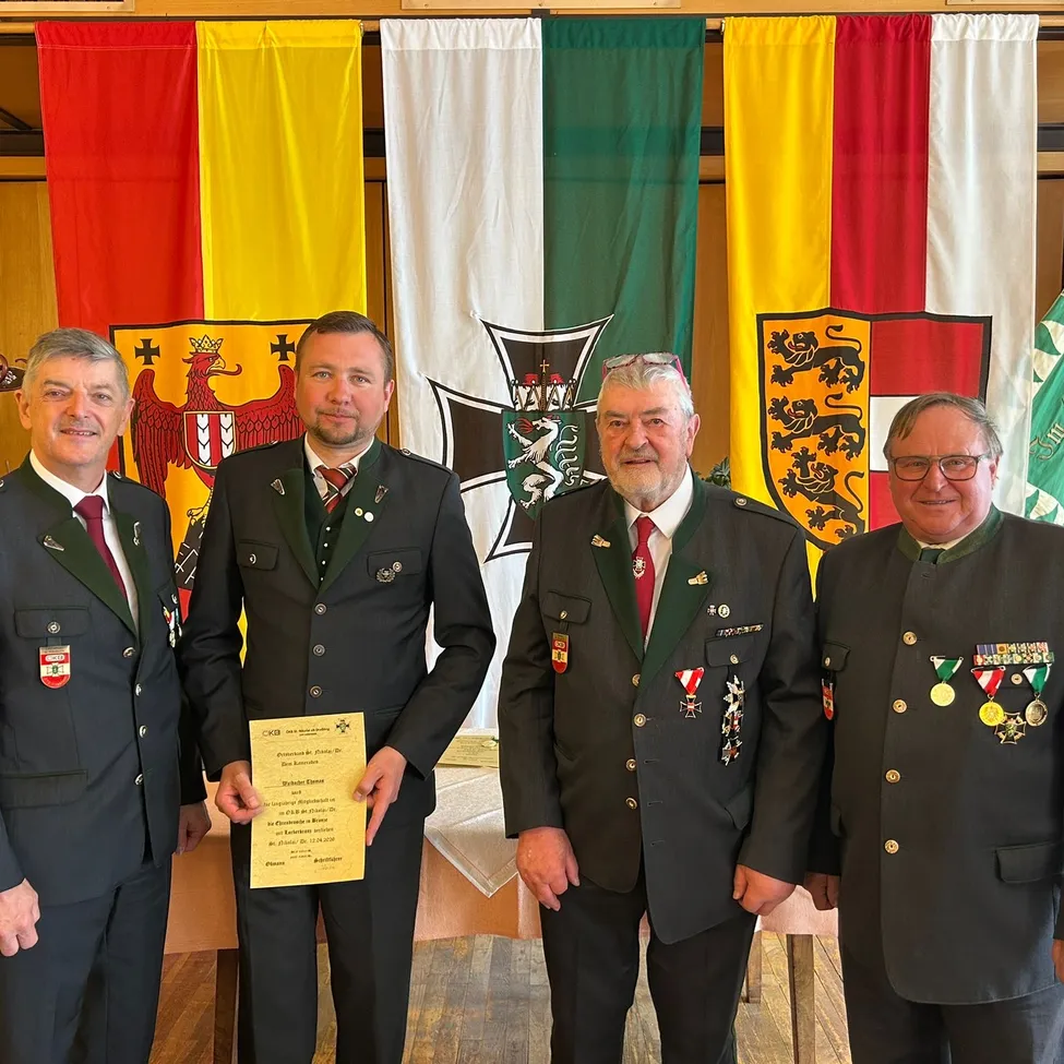 Vier Männer in Uniformen mit Medaillen stehen vor Fahnen. Der Mann in der Mitte hält ein Zertifikat.