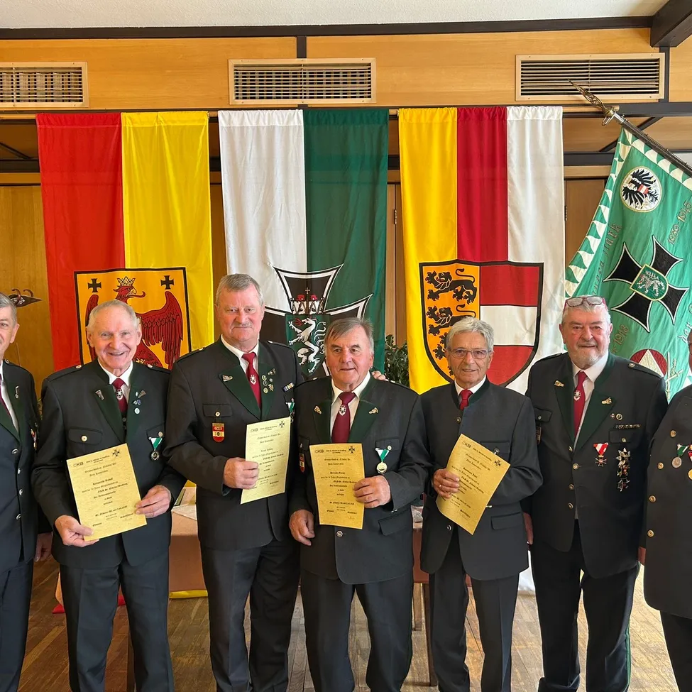 Eine Gruppe von Männern in Uniformen steht vor Flaggen und hält Urkunden. Sie scheinen eine besondere Leistung zu feiern.