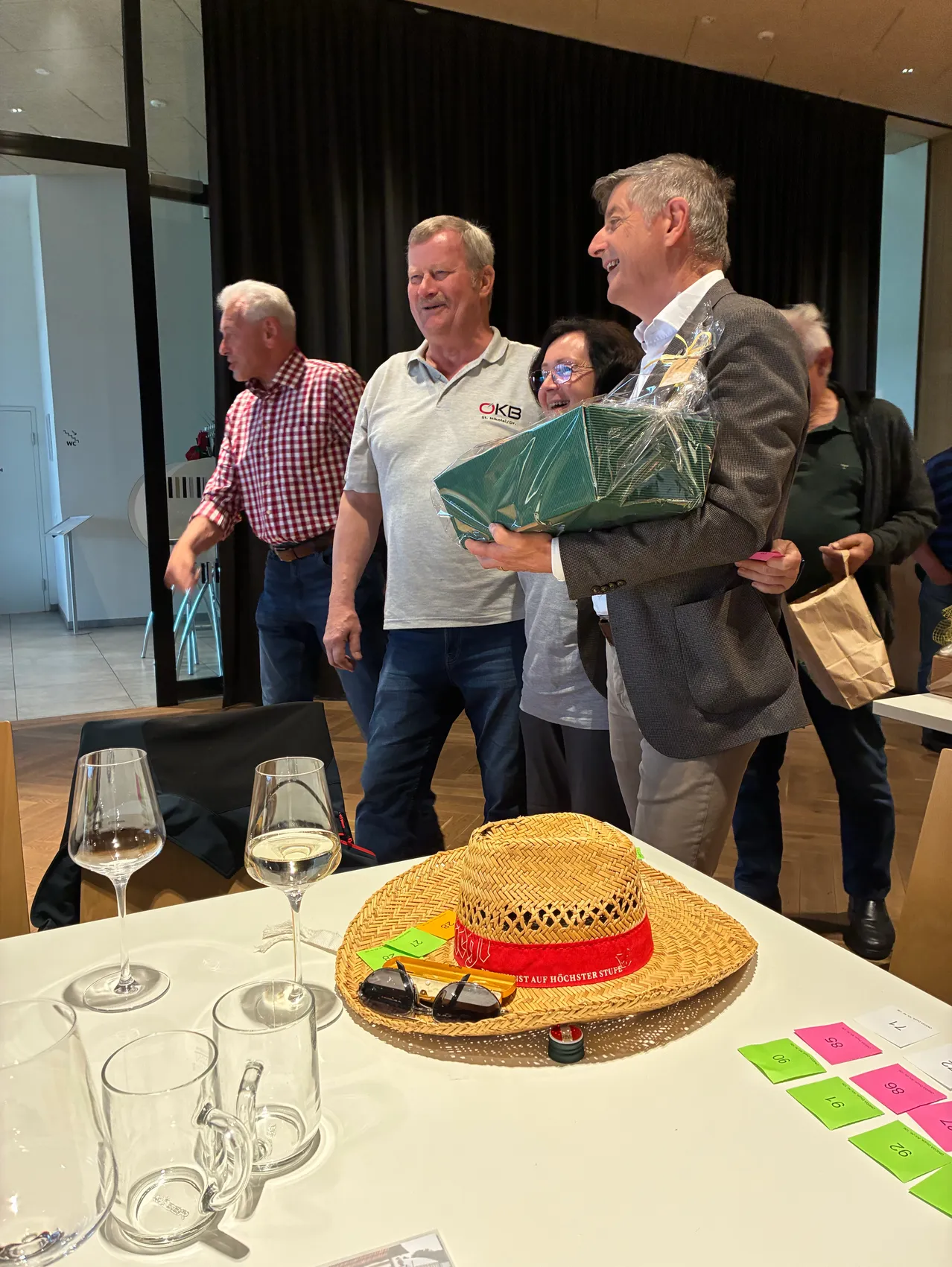 Mehrere Menschen stehen in einem Raum mit einem Tisch. Ein Mann hält ein eingewickeltes Geschenk und lächelt, ein anderer trägt einen Hut mit rotem Text. Ein Tisch hat Gläser, einen Strohhut und Papiere mit Zahlen.