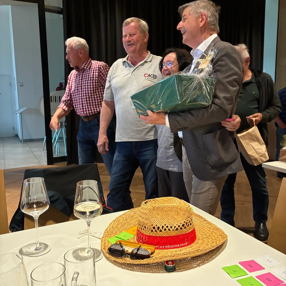 Mehrere Menschen stehen in einem Raum mit einem Tisch. Ein Mann hält ein eingewickeltes Geschenk und lächelt, ein anderer trägt einen Hut mit rotem Text. Ein Tisch hat Gläser, einen Strohhut und Papiere mit Zahlen.