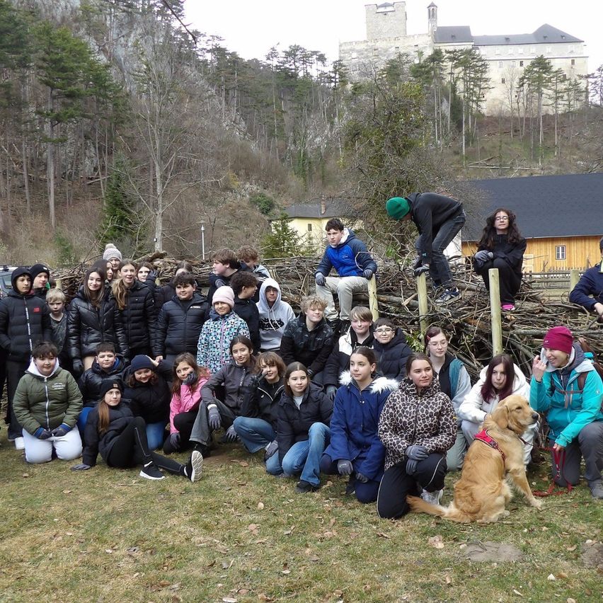 Eine Gruppe von Menschen versammelt sich in einem Park und trägt Winterkleidung und einige Mützen. Einige knien, andere stehen auf einem Haufen von Stöcken, und ein Hund sitzt auf dem Boden. Dahinter befindet sich ein Schloss und ein Haus.