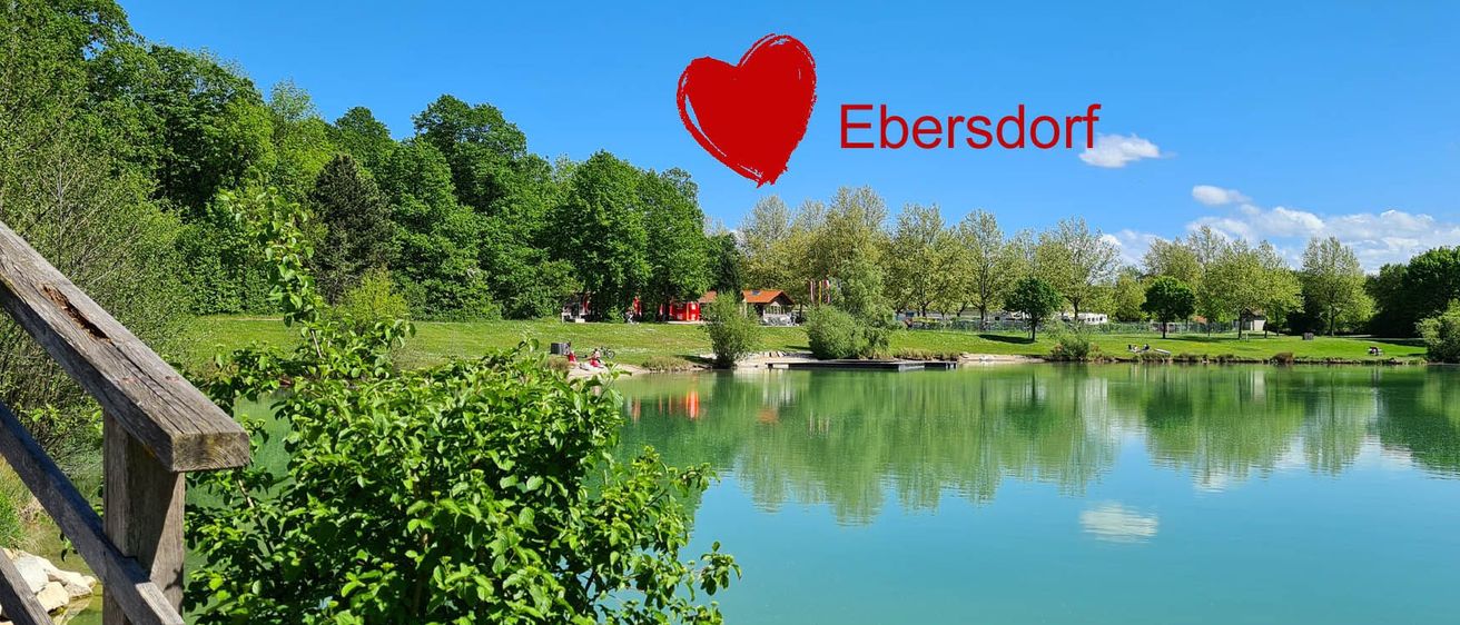 Eine friedliche Landschaft mit klarem blauem Himmel, einem See, der grüne Bäume widerspiegelt, und einem roten Herzsymbol mit der Aufschrift 'Ebersdorf'.
