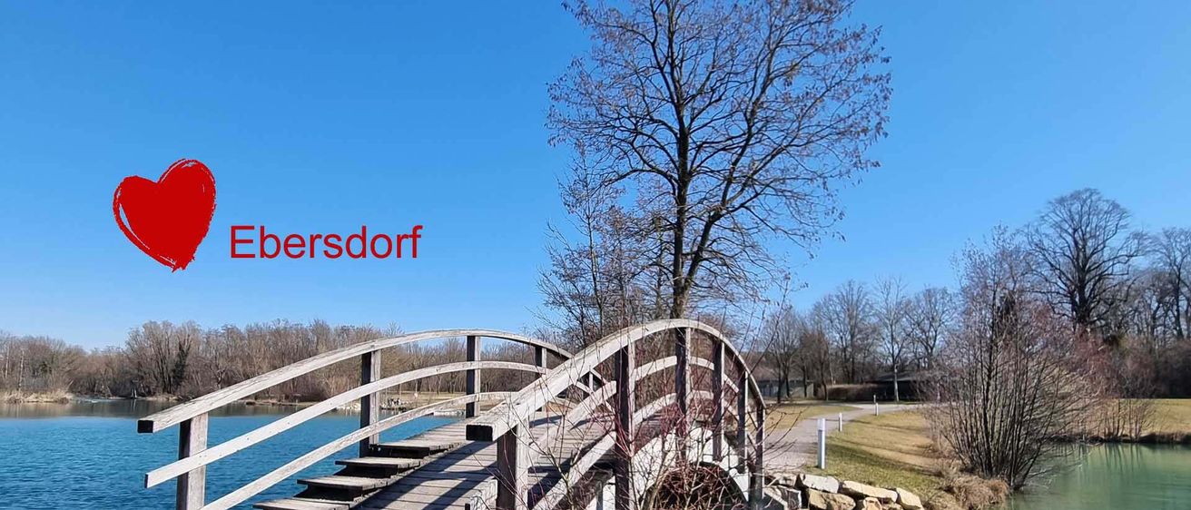 Eine Holzbrücke überspannt einen ruhigen See, umgeben von Bäumen mit wenigen Blättern, unter einem klaren blauen Himmel. Das Wort 'bersddorf' ist prominent in Rot über der Brücke angebracht.