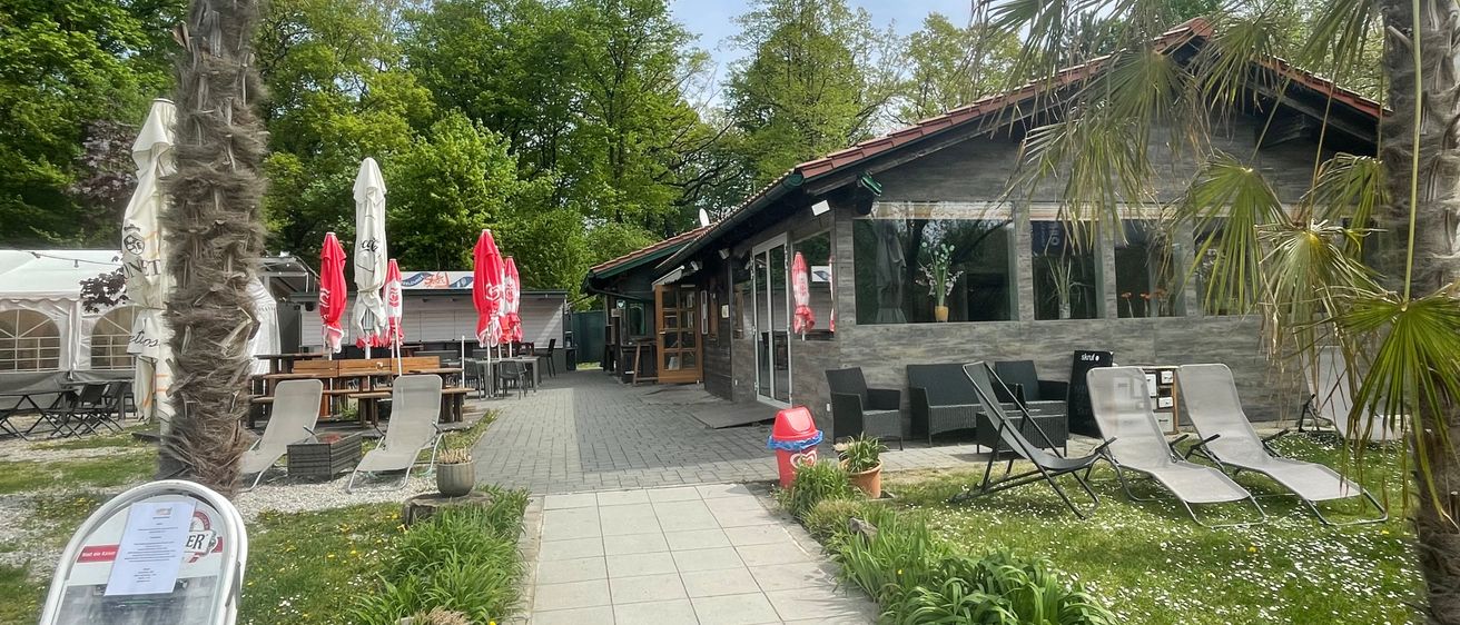 Outdoor-Café mit roten Sonnenschirmen, Liegestühlen und einem Weg in einem Grasbereich mit Bäumen im Hintergrund.