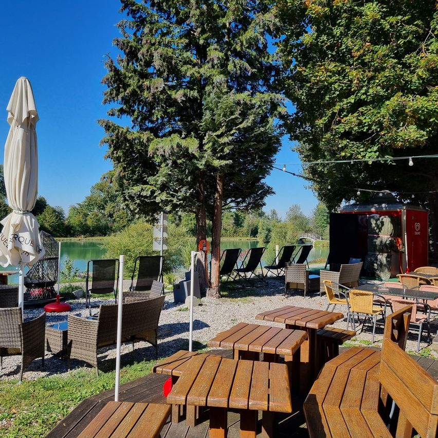 Eine Außensitzgelegenheit in einem Restaurant mit Tischen und Stühlen unter Sonnenschirmen, neben einem See mit klarem blauem Himmel und Bäumen im Hintergrund.