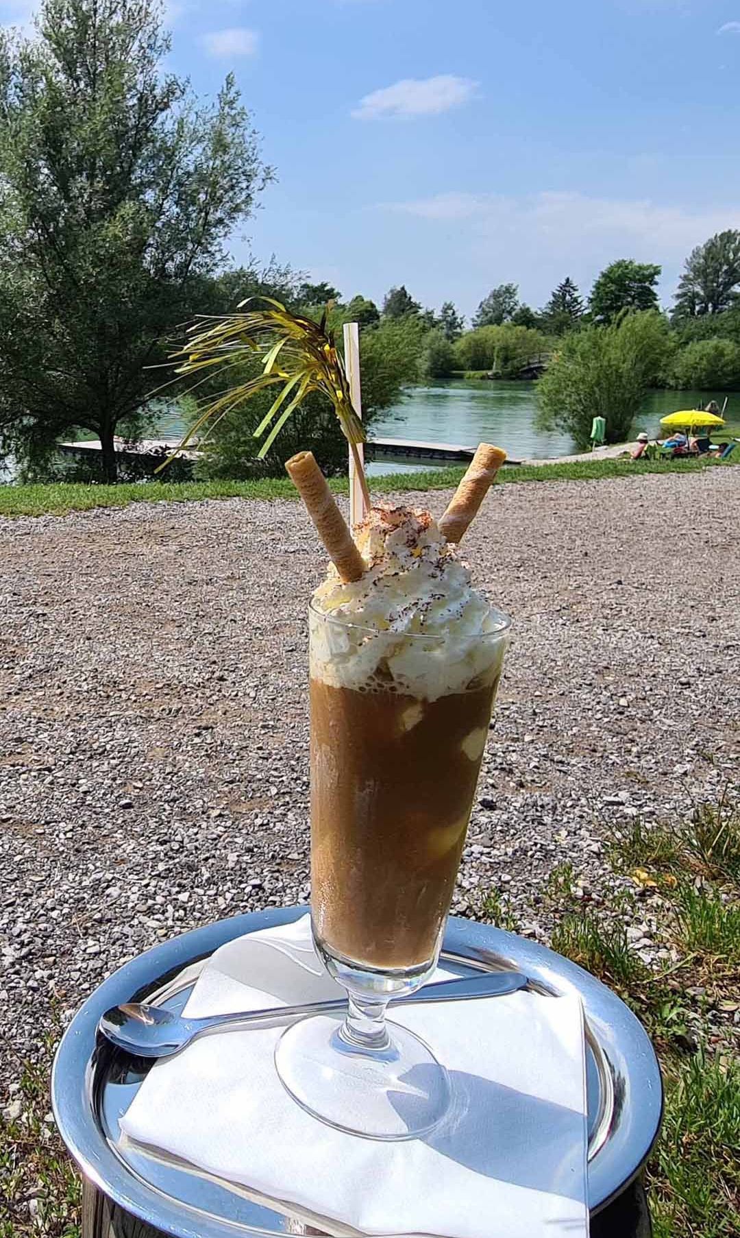 Ein hohes Glas mit einem Schokoladenmilchshake mit Schlagsahne und zwei Eisbechern darauf. Es steht auf einem Tablett in der Nähe eines Sees, wo Leute unter einem Regenschirm sitzen.