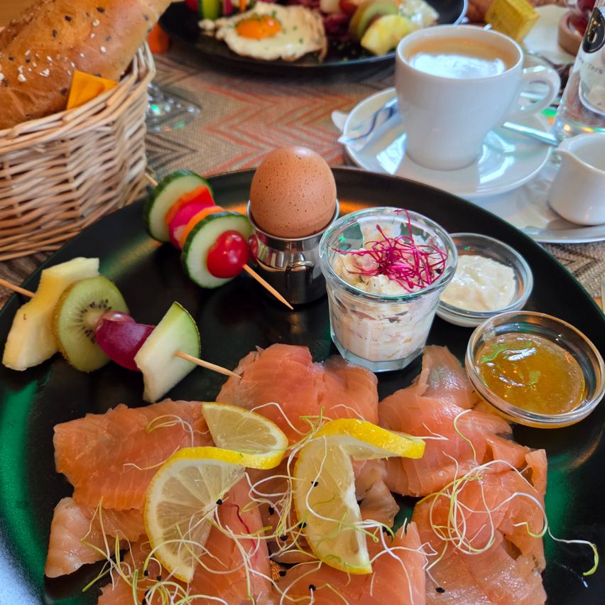 Ein Frühstücksteller mit Lachs, Gurken- und Obstspießen, Zitronenscheiben und Eiern auf einer Untertasse. Ein Korb mit Brot und eine Tasse Kaffee auf dem Tisch.