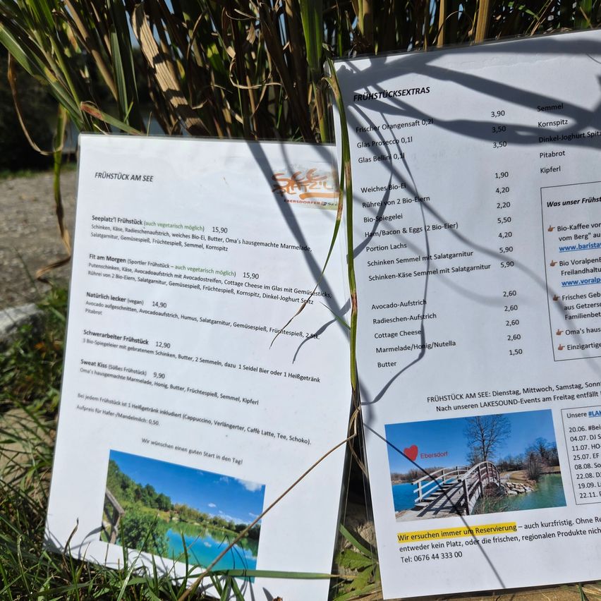 Zwei Frühstücksmenüs liegen auf Gras. Das erste Menü listet verschiedene Frühstücksartikel mit Preisen und Ernährungsoptionen auf. Das zweite Menü listet Frühstücksartikel mit Preisen auf, einschließlich Artikel wie 'Frischer Orangensaft' und 'Marmelade/Honig/Nussella'.