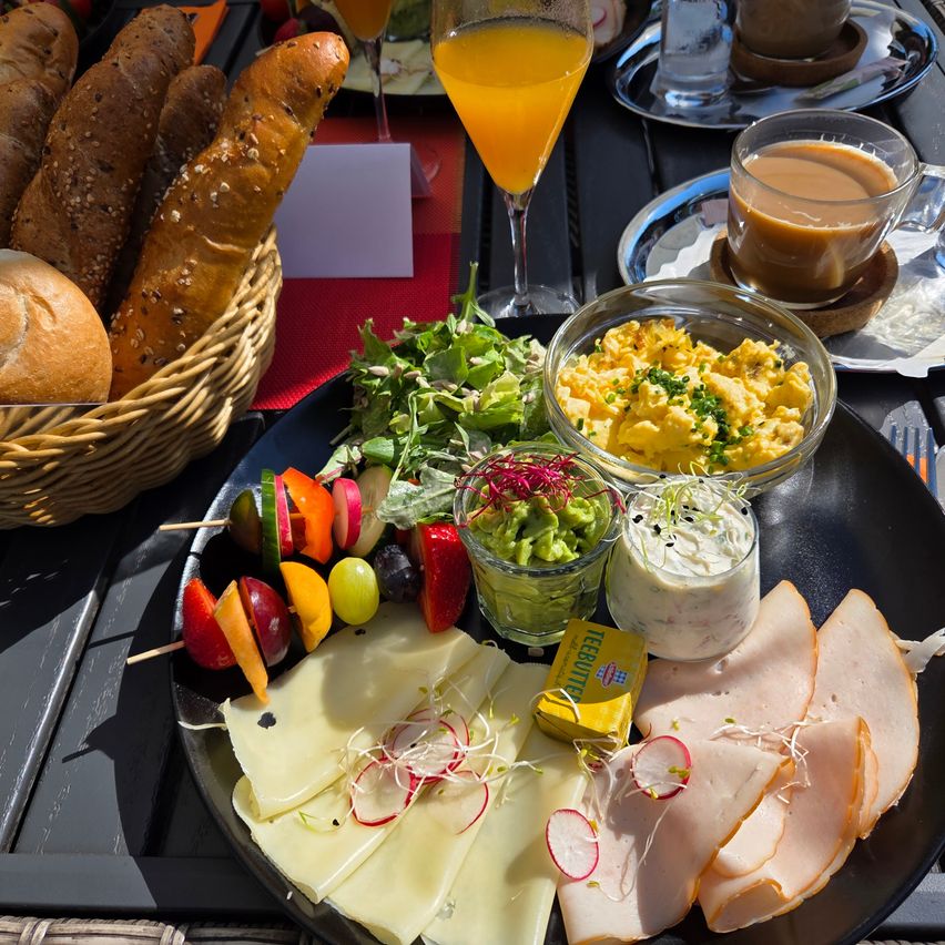 Ein Frühstücksbuffet auf einem Tisch besteht aus einer großen Platte mit Käse, Fleisch, Radieschen und Salat. Ein Korb mit Brot und ein Glas Orangensaft stehen in der Nähe. Zwei Tassen Kaffee und ein Glas Wasser sind auf dem Tisch.