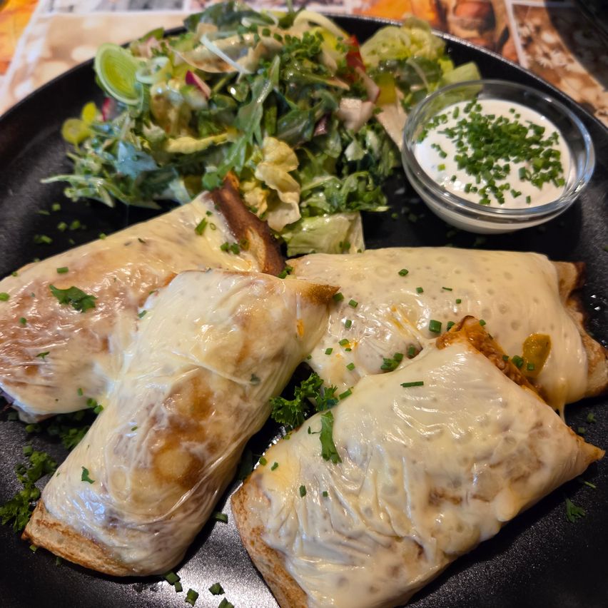 A black plate with several wrapped pastries, garnished with parsley and served with a salad and a small bowl of creamy dip.