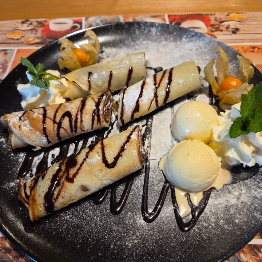 A plate with three rolled pastries, each drizzled with chocolate, accompanied by whipped cream and ice cream, garnished with mint leaves and powdered sugar.