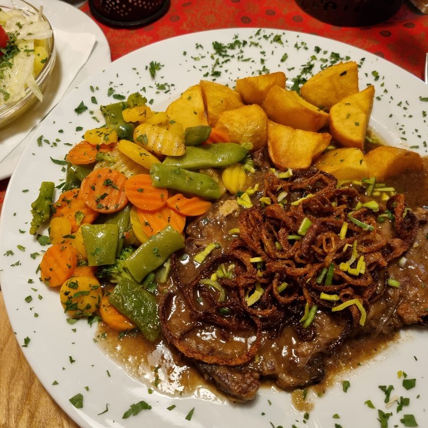 Ein weißer Teller mit einem Fleischgericht, bedeckt mit Soße, serviert mit gebratenen Zwiebeln, Karotten, grünen Bohnen und Kartoffeln, garniert mit Kräutern auf einem roten Tischtuch.