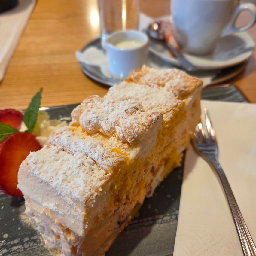 Ein Stück Kuchen mit Puderzucker auf einem Teller mit einer Gabel und einer Erdbeere. Eine Tasse und Untertasse stehen auf einem Holztisch.