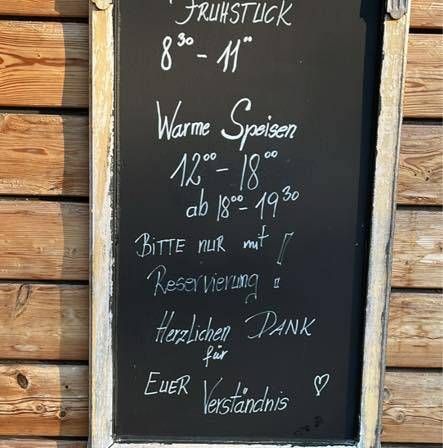 Eine Holzwand hält ein gerahmtes Tafel mit weißer Schrift. Es steht 'Fruhstuck 8:30-11' und 'Warme Speisen 12-18'. Es gibt eine Nachricht 'BITTE NUR MIT RESERVIERUNG'. Darunter steht 'Herzlichen Dank für EUER Verständnis'. Ein Emoji ist unten rechts.