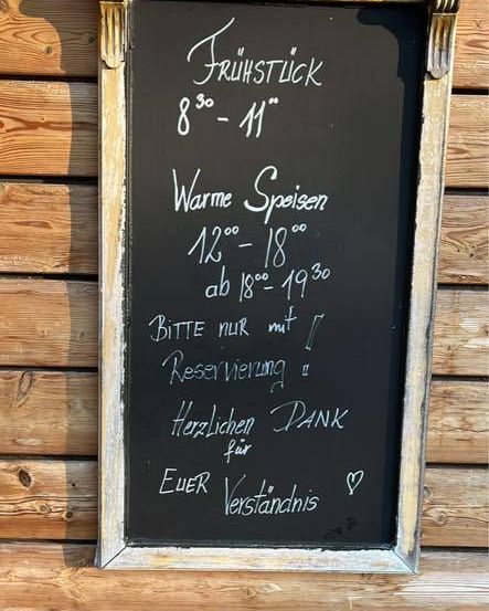 Eine Holzwand hält ein gerahmtes Tafel mit weißer Schrift. Es steht 'Fruhstuck 8:30-11' und 'Warme Speisen 12-18'. Es gibt eine Nachricht 'BITTE NUR MIT RESERVIERUNG'. Darunter steht 'Herzlichen Dank für EUER Verständnis'. Ein Emoji ist unten rechts.