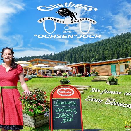 Eine Frau in einem roten Kleid steht vor einem Chalet mit einem Willkommensschild. Hinter ihr steht ein Gebäude mit mehreren Sonnenschirmen und Tischen auf einer Wiese. Im Hintergrund befinden sich Bäume und Berge.