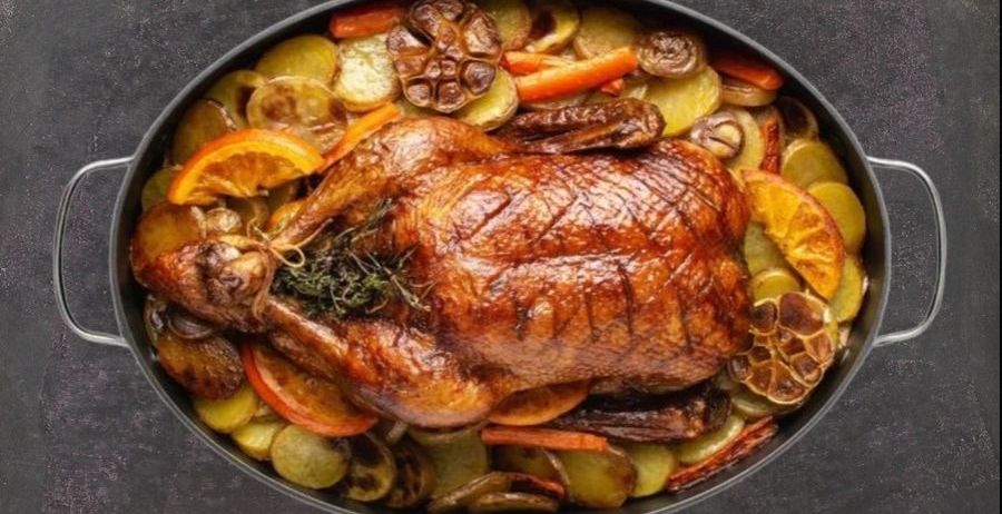 Eine gebratene Ente liegt in einer Bratpfanne mit verschiedenen Gemüsesorten, einschließlich Karotten, Kartoffeln und Knoblauch. Die Ente ist mit Kräutern garniert.