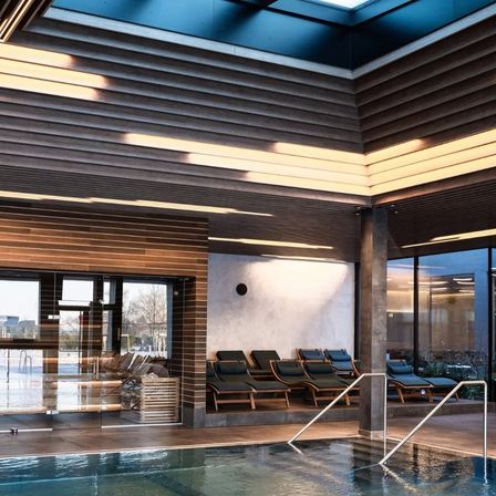 Ein Hallenbad mit einer Holzdecke und Liegestühlen an der Wand. Der Pool ist sauber und hat eine Glasbarriere.