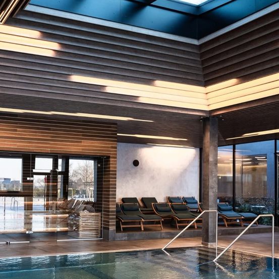 Ein Hallenbad mit einer Holzdecke und Liegestühlen an der Wand. Der Pool ist sauber und hat eine Glasbarriere.
