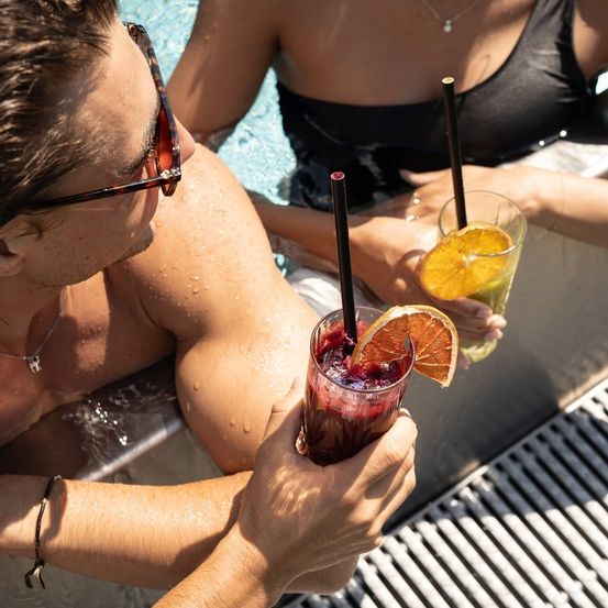 Ein Mann und eine Frau im Pool, jeder hält ein Getränk mit Strohhalm und Orangenscheibe. Der Mann trägt eine Sonnenbrille und eine Halskette.