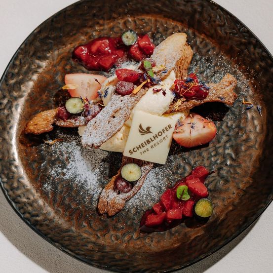 Ein Gourmet-Dessertteller mit verschiedenen Früchten wie Erdbeeren, Himbeeren und Wassermelonen auf einer Schicht Schlagsahne, garniert mit Puderzucker und essbaren Blüten.