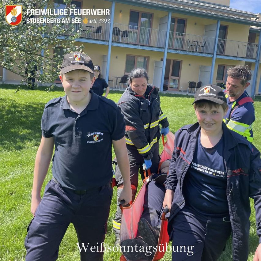 Drei Personen in Feuerwehruniformen üben an einem sonnigen Tag vor einem Gebäude eine Notfallrettung.