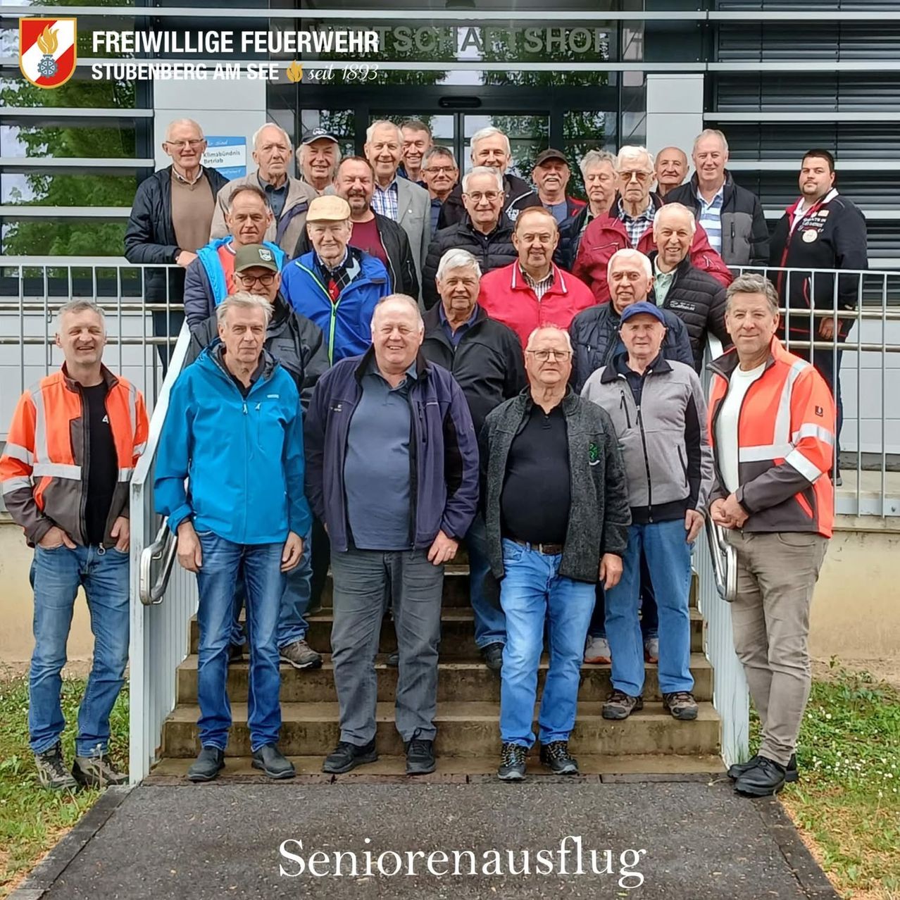 Eine Gruppe von Senioren der Freiwilligen Feuerwehr Stubenerberg am See steht auf einer Treppe vor einem Gebäude und posiert für ein Foto. Die Gruppe trägt Jacken, Hosen und Schuhe.
