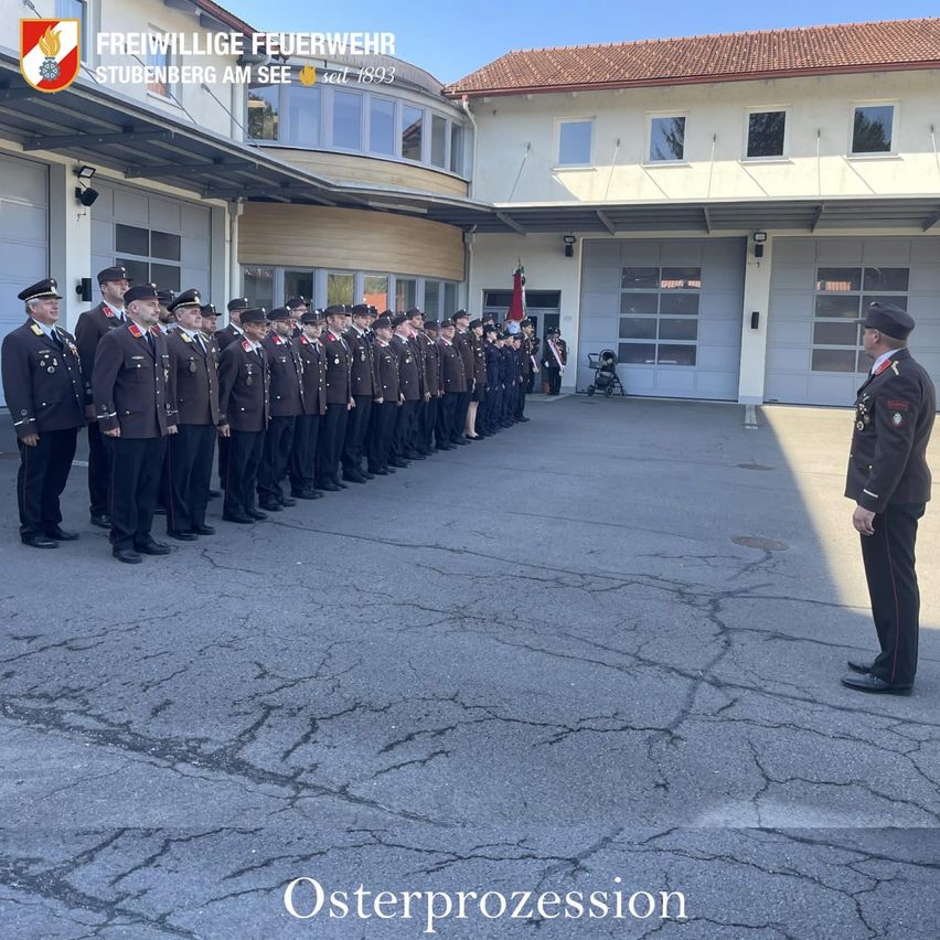 Eine Gruppe von Feuerwehrleuten in Uniform steht in einer Reihe, möglicherweise bei einer Osterprozession, vor einem Gebäude mit gefliestem Dach und einem Schild, das 'Freiwillige Feuerwehr Stubenber AM See seit 1893' besagt.