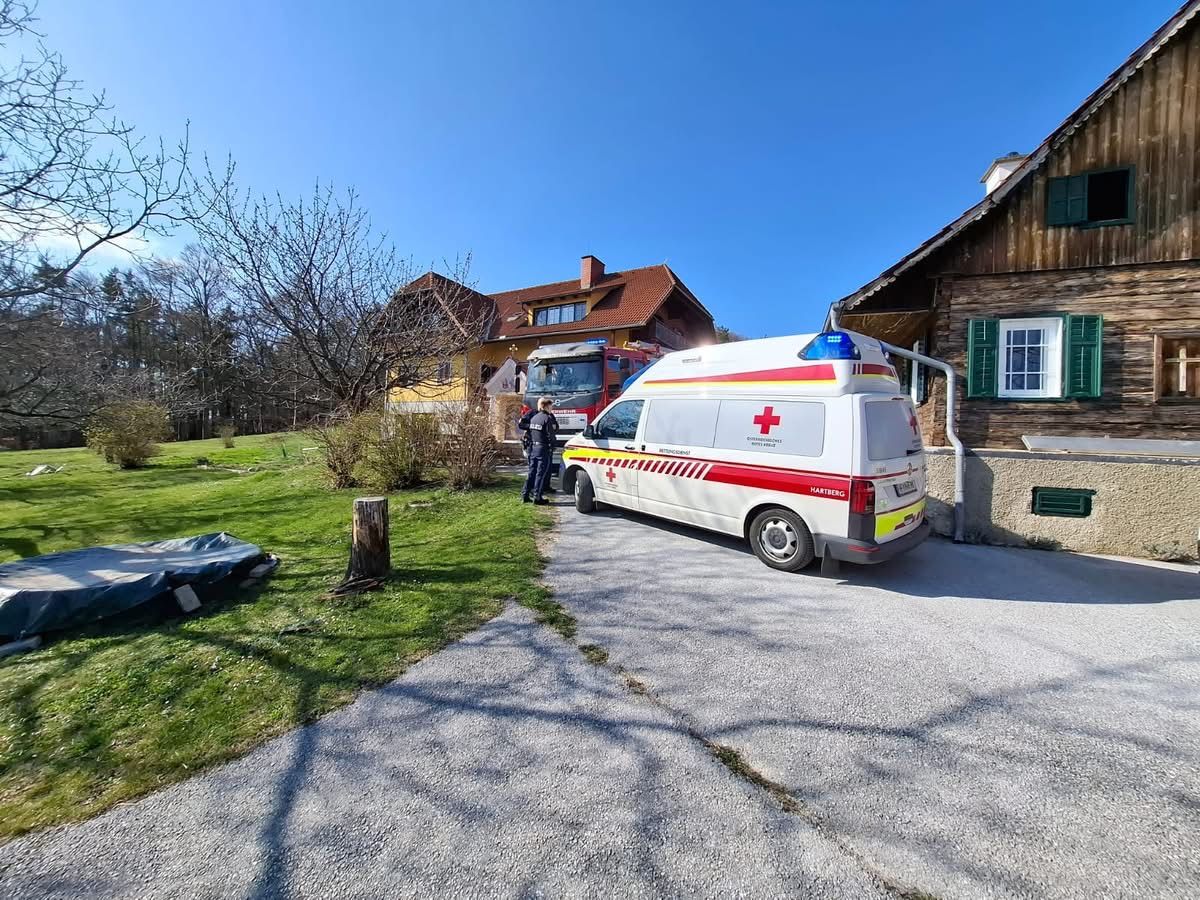 Ein Krankenwagen ist vor einem Haus geparkt. Zwei Personen stehen in der Nähe des Krankenwagens, und ein Feuerwehrauto ist dahinter geparkt.