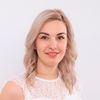 Eine lächelnde Frau mit blonden gewellten Haaren, die ein weißes Spitzenoberteil trägt. Sie schaut fröhlich nach vorne.